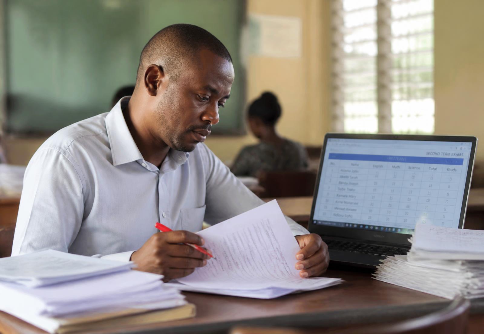 A teacher entering exam marks beside a stack of marked papers.
